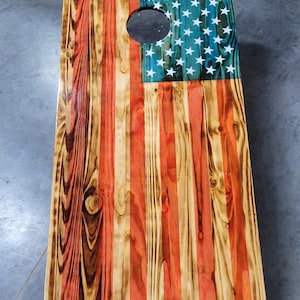 May include: A wooden American flag cornhole board with a red, white, and blue design. The board has a hole in the center for tossing bean bags.