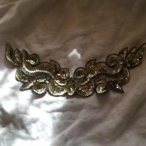 May include: A gold sequined applique with a swirling design. The applique is on a white fabric background.