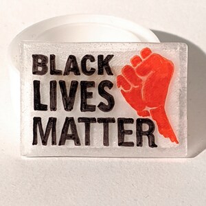 Könnte beinhalten: Ein weißes rechteckiges Schild mit den Worten "Black Lives Matter" in schwarzer Schrift. Auf der rechten Seite des Schildes ist eine rote geballte Faust abgebildet.