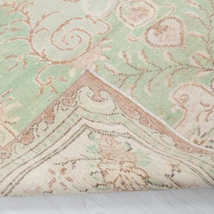 May include: A vintage-style rug with a distressed appearance. The rug features a light green field with faded brown and beige floral patterns. The border is a darker brown with intricate designs, and a portion of the rug is folded over.