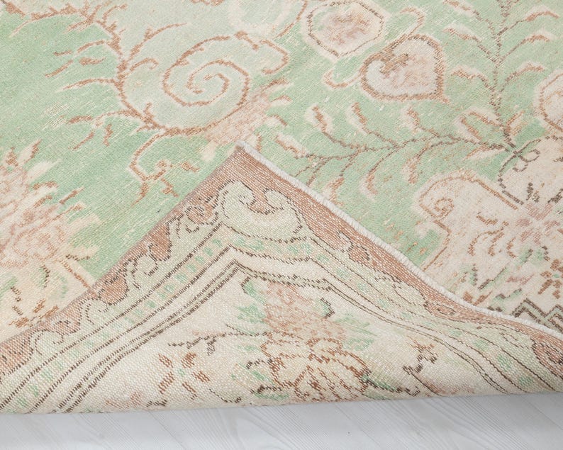 May include: A vintage-style rug with a distressed appearance. The rug features a light green field with faded brown and beige floral patterns. The border is a darker brown with intricate designs, and a portion of the rug is folded over.