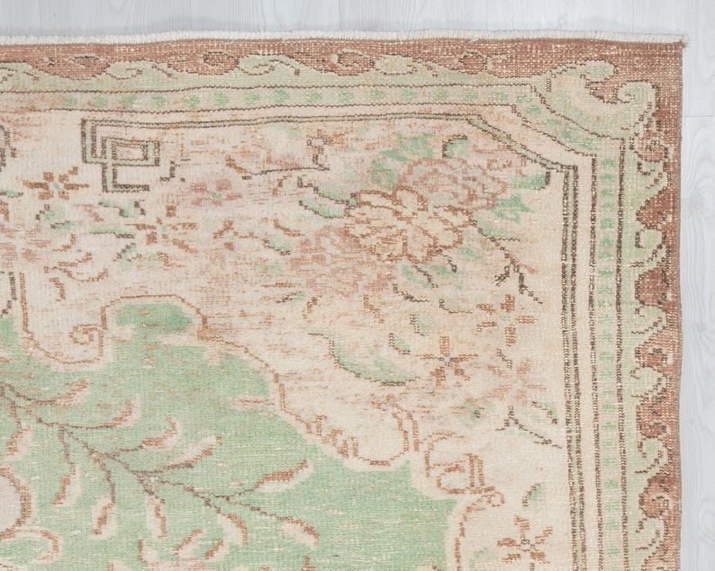 May include: Close-up of a vintage rug with a distressed, faded appearance. The rug features a cream-colored central field with floral and geometric patterns in shades of green, brown, and peach. The border is a darker brown with a repeating design.