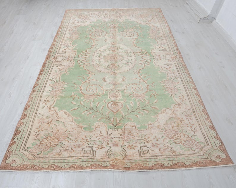 May include: A large, rectangular area rug with an intricate floral pattern. The rug features a central field of light green, surrounded by a border of beige and brown. Floral and vine motifs are spread across the surface.