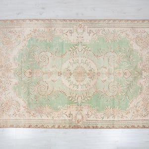 May include: Rectangular area rug with a central green field and a beige border. The rug features an intricate floral pattern in shades of beige, brown, and green. The rug's design is symmetrical, with a central medallion and corner motifs.