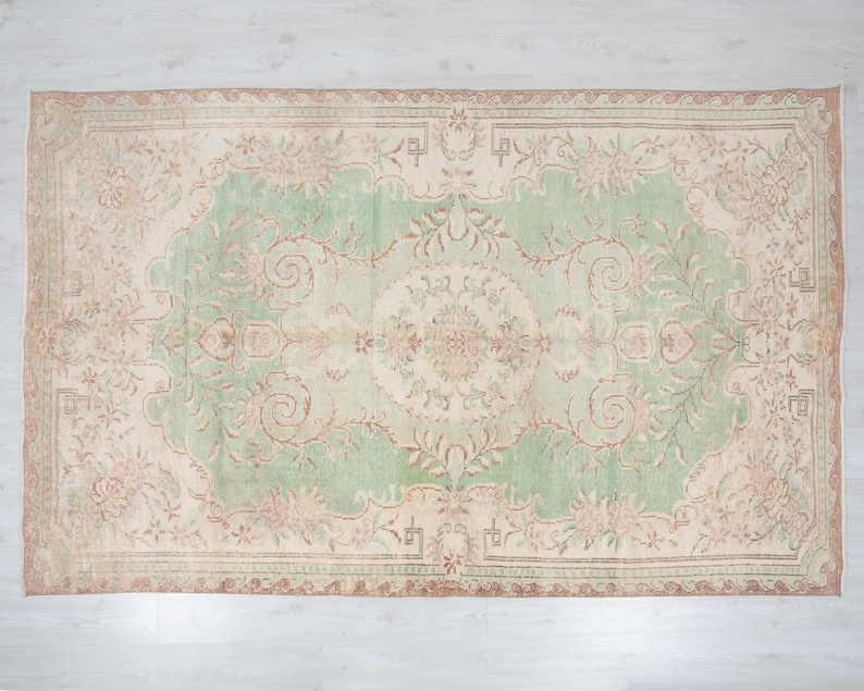 May include: Rectangular area rug with a central green field and a beige border. The rug features an intricate floral pattern in shades of beige, brown, and green. The rug's design is symmetrical, with a central medallion and corner motifs.