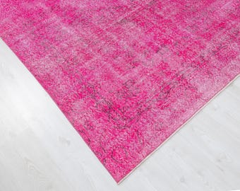 May include: A close-up of a vibrant pink rug with a distressed, vintage appearance. The rug's texture is visible, with variations in the pink hue and darker areas. The rug is on a white surface.