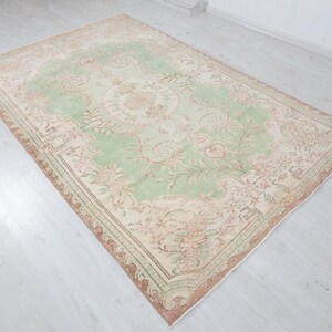 May include: A rectangular, vintage-style rug with a central light green field featuring floral and vine patterns. The border is a mix of beige, brown, and pink tones. The rug is placed on a light-colored wooden floor.