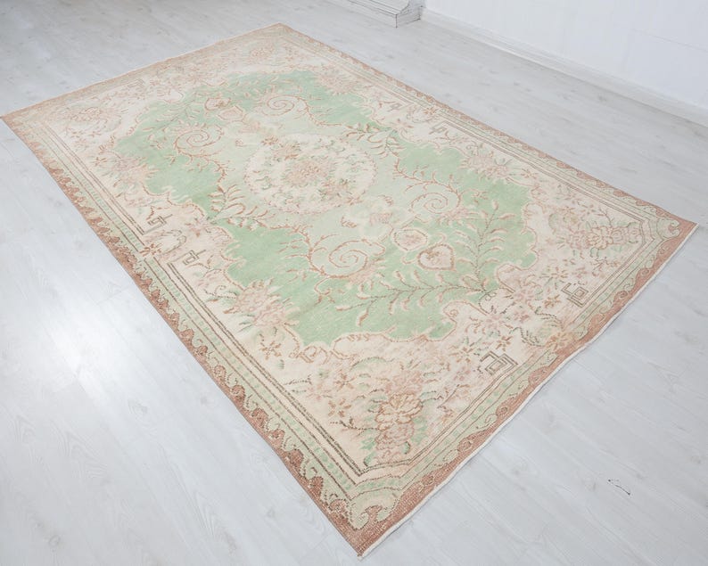 May include: A rectangular, vintage-style rug with a central light green field featuring floral and vine patterns. The border is a mix of beige, brown, and pink tones. The rug is placed on a light-colored wooden floor.