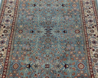 May include: A rectangular area rug featuring a light blue field with a dense, symmetrical floral pattern in shades of cream, dark red, and navy blue. The rug is framed by a wide border with intricate botanical motifs on a cream background.