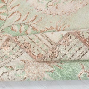 May include: Close-up of a folded, vintage-style rug. The rug features a faded floral pattern in shades of green, beige, and brown. The edges are detailed with a border design. The rug's texture appears soft and worn, suggesting it is a well-loved piece.