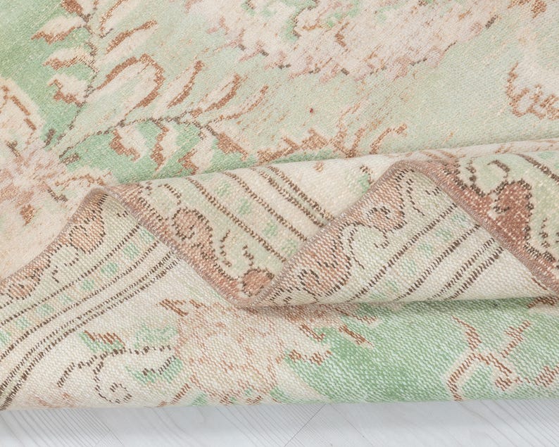 May include: Close-up of a folded, vintage-style rug. The rug features a faded floral pattern in shades of green, beige, and brown. The edges are detailed with a border design. The rug's texture appears soft and worn, suggesting it is a well-loved piece.