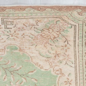 May include: Close-up of a vintage rug with a distressed, faded appearance. The rug features a cream-colored central field with floral and geometric patterns in shades of green, brown, and peach. The border is a darker brown with a repeating design.