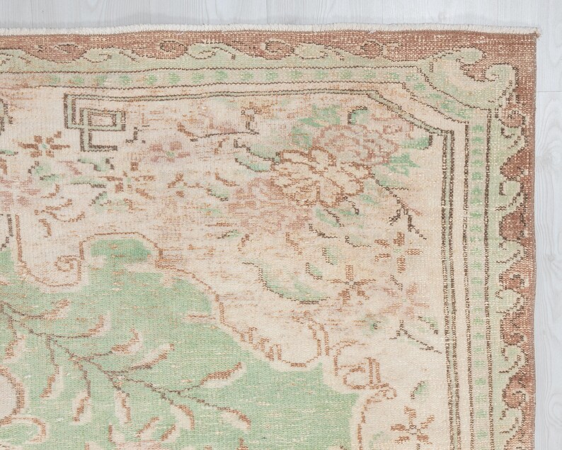 May include: Close-up of a vintage rug with a distressed, faded appearance. The rug features a cream-colored central field with floral and geometric patterns in shades of green, brown, and peach. The border is a darker brown with a repeating design.