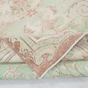May include: A rolled-up, vintage-style rug with a faded, distressed appearance. The rug features a cream-colored background with a repeating pattern in shades of green and brown. The border is a darker brown with intricate designs.