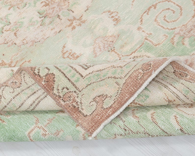 May include: A rolled-up, vintage-style rug with a faded, distressed appearance. The rug features a cream-colored background with a repeating pattern in shades of green and brown. The border is a darker brown with intricate designs.
