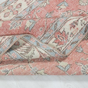 May include: Close-up of a folded vintage rug with a faded pink and blue floral pattern. The rug has a distressed look and is made of a soft, woven material.