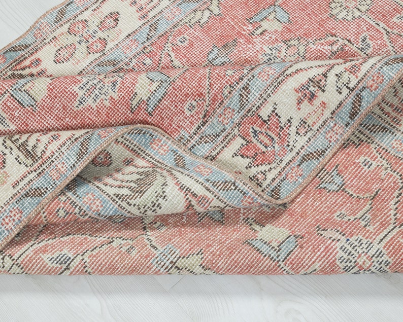 May include: Close-up of a folded vintage rug with a faded pink and blue floral pattern. The rug has a distressed look and is made of a soft, woven material.