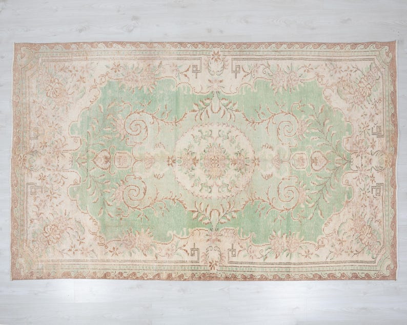 May include: Rectangular area rug with a central green field and a beige border. The rug features an intricate floral pattern in shades of beige, brown, and green. The rug's design is symmetrical, with a central medallion and corner accents.