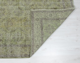 May include: A distressed, vintage-style rug with a faded green and brown color palette. The rug features an intricate floral pattern and a dark brown border. The corner of the rug is folded over, revealing the underside and the white floor.