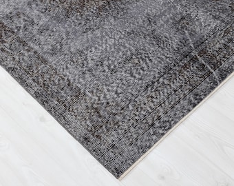 Op de afbeelding: Een hoekweergave van een versleten, rechthoekig tapijt met een donkergrijs en bruin patroon. Het tapijt heeft een rand en ligt op een witte houten vloer. Het ontwerp van het tapijt heeft een vintage, vervaagde uitstraling.