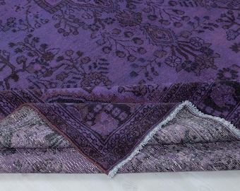 May include: A deep purple area rug featuring a dark, intricate floral and geometric pattern. The rug is folded over to reveal a textured underside with a visible fringe edge, showcasing a consistent color palette of violet and black throughout.