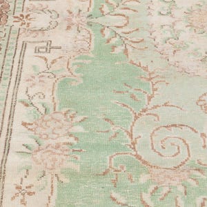 May include: Close-up of a vintage rug with a faded floral pattern. The rug features a central field of light green, bordered by a cream-colored frame and accents of brown. The design includes stylized flowers and scrolling vines.