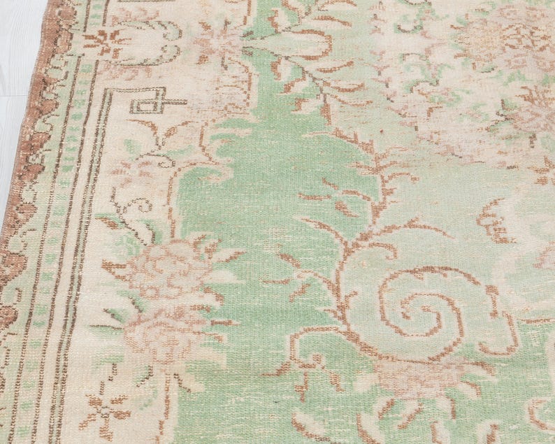 May include: Close-up of a vintage rug with a faded floral pattern. The rug features a central field of light green, bordered by a cream-colored frame and accents of brown. The design includes stylized flowers and scrolling vines.