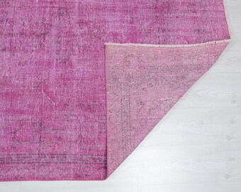 May include: A distressed, vintage-style rug in shades of fuchsia and pink. The rug has a faded, textured appearance with darker areas, and a portion is folded over, revealing the underside. The rug is placed on a white surface.