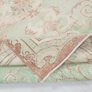 May include: A rolled-up, vintage-style rug with a faded, distressed appearance. The rug features a cream-colored background with a repeating pattern in shades of green and brown. The border is a darker brown with intricate designs.