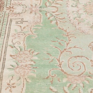 May include: Close-up of a vintage rug with a faded floral design. The rug features a central field of light green with beige and brown floral patterns. The border is a darker brown with a repeating geometric pattern. The rug has a distressed, antique look.