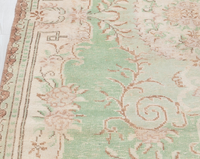 May include: Close-up of a vintage rug with a faded floral design. The rug features a central field of light green with beige and brown floral patterns. The border is a darker brown with a repeating geometric pattern. The rug has a distressed, antique look.