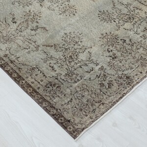 May include: A rectangular area rug featuring a faded light gray and beige background with a repeating dark brown floral pattern. The rug has a wide, dark brown border with intricate botanical motifs and a visible woven texture throughout the surface.