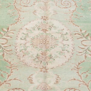 May include: A close-up of a vintage rug with a faded floral pattern. The rug features a central medallion in cream and tan, surrounded by a light green field with brown floral accents. The overall design has a distressed, antique look.