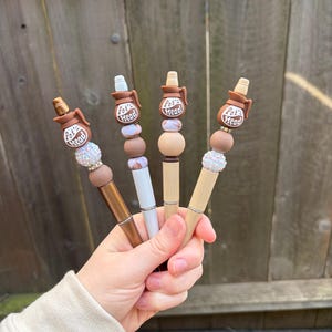 May include: Four pens with decorative tops are held in a hand. Each pen features a brown ceramic pot design with the text "Pot's Head". The pens have various bead accents in neutral tones and metallic finishes.