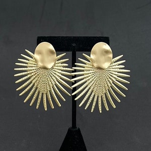 Puede incluir: Un par de pendientes dorados. Cada pendiente presenta una parte superior circular texturizada con un diseño de rayos de sol. Los pendientes se exhiben en un soporte negro sobre un fondo oscuro.