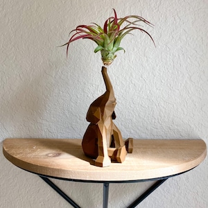 May include: A brown, geometric elephant figurine holding an air plant with green and red leaves. The elephant is sitting, with its trunk raised to support the plant. The figurine is on a wooden shelf.