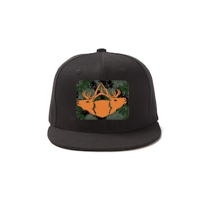 Puede incluir: Gorra de béisbol negra con un parche verde y naranja con tres cabezas de ciervo. El parche tiene un patrón de camuflaje.