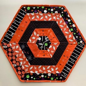May include: A hexagonal table runner with a Halloween theme. The runner is made of orange, black, and white fabric with a pattern of ghosts, pumpkins, and stars. The edges of the runner are trimmed with a black and white polka dot fabric.