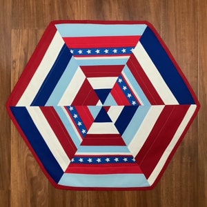 May include: A hexagonal table runner with a geometric pattern in red, white, and blue. The pattern features stripes and stars.