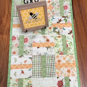 May include: A rectangular fabric table runner featuring a patchwork design of floral, hummingbird, and butterfly patterns in shades of green, orange, and white. A square wooden sign with a yellow honeycomb background and the text BEE HAPPY.