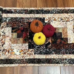 Peut inclure: Un chemin de table matelassé rectangulaire présentant un motif en patchwork avec des imprimés floraux et abstraits noirs, marron, crème et beige. Trois petites citrouilles en tissu texturé orange, rouge foncé et jaune sont posées au centre.