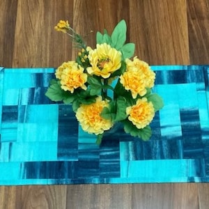 May include: A turquoise and blue patchwork table runner with a floral arrangement of yellow flowers in the center.