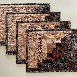 May include: A set of four rectangular quilted placemats featuring a log cabin pattern. The fabric displays a variety of floral and leaf prints in shades of brown, tan, cream, and black, with dark borders framing each individual quilted piece.