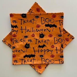 May include: Two square fabric coasters featuring an orange background with a repeating black print of bats, spiders, cats, and pumpkins. The text reads Trick or Treat, Boo, and Happy Halloween in a handwritten style font across the fabric surface.