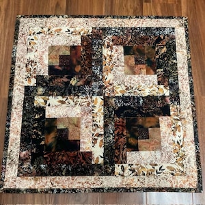 Peut inclure: Un quilt carré présentant un motif de blocs en cabane de bûcheron dans des tons de noir, marron, crème et beige. Le design intègre divers imprimés floraux et de feuilles, avec une large bordure encadrant les quatre blocs centraux.