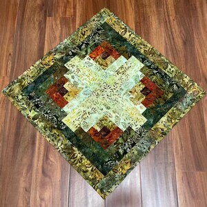 May include: A square quilted fabric mat featuring a log cabin pattern with a central light cream section surrounded by concentric layers of fabric in shades of dark green, olive, brown, and rust orange, all decorated with a repeating leaf print.