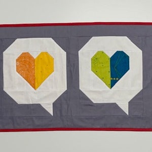 May include: A gray fabric banner with a red border features four white speech bubbles with colorful hearts inside. The hearts are red and orange, orange and yellow, green and blue, and pink and purple.