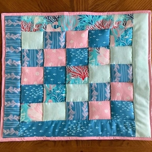 May include: A pink and blue patchwork quilt with a sea life theme. The quilt is made up of squares of different fabrics, each with a different design. The designs include fish, coral, and seaweed. The quilt is edged with a pink border.