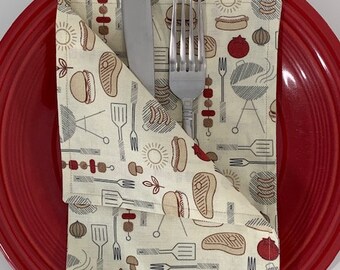 May include: A bright red dinner plate holding a folded cream-colored cloth napkin with a repeating pattern of barbecue-themed illustrations including grills, spatulas, forks, skewers, steaks, hot dogs, burgers, onions, and sun icons.