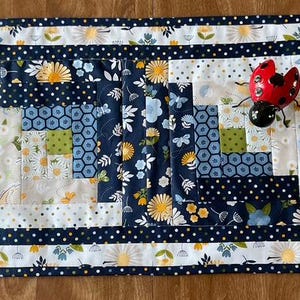 May include: A rectangular quilted fabric mat featuring a log cabin pattern with navy blue, white, yellow, and light blue floral and geometric prints. A small red and black ladybug figurine is positioned on the top right corner of the mat.